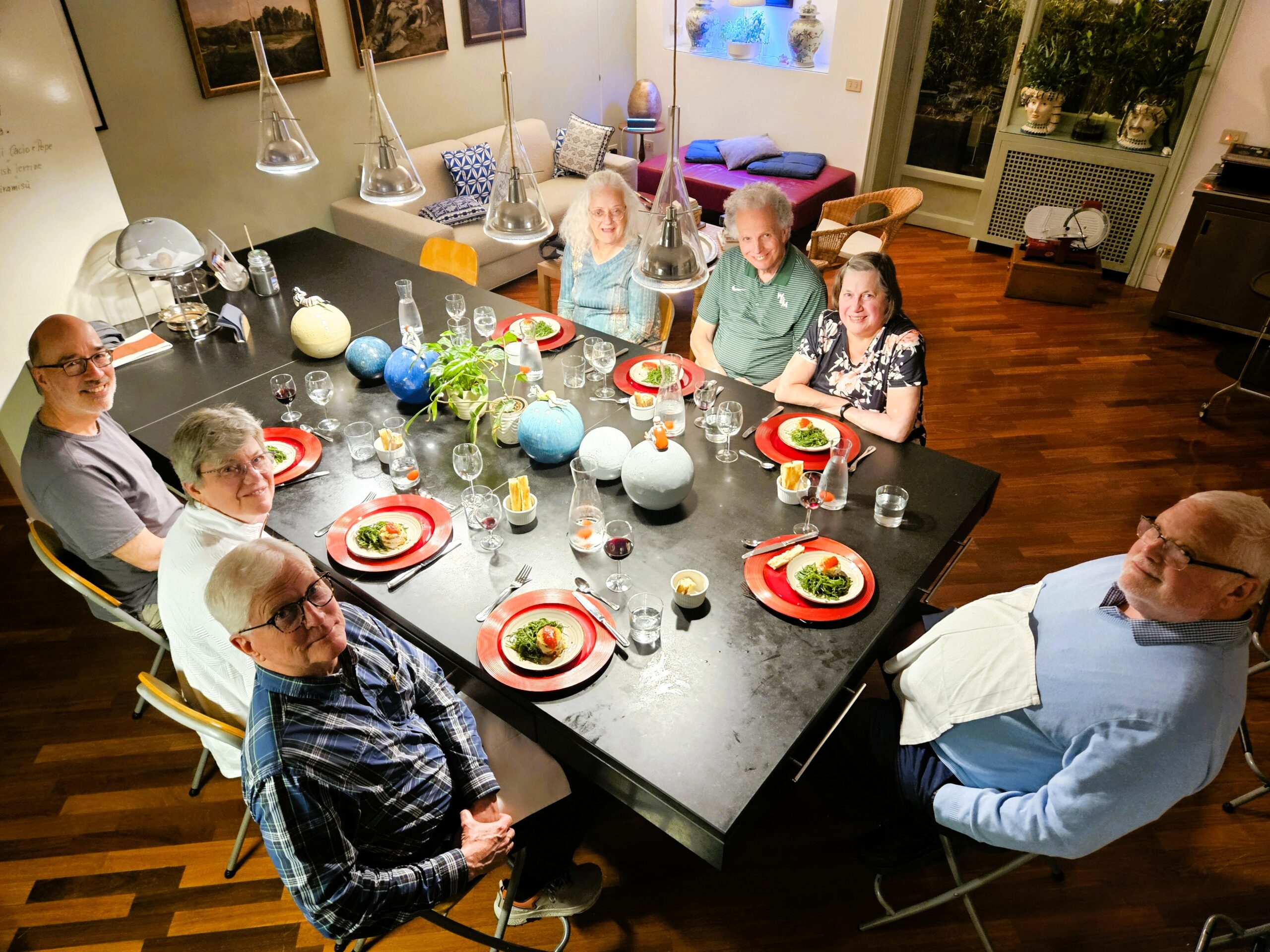 A group of smiling seniors sits around a long dining table with red plates, enjoying a home-cooked meal in a warmly lit room.