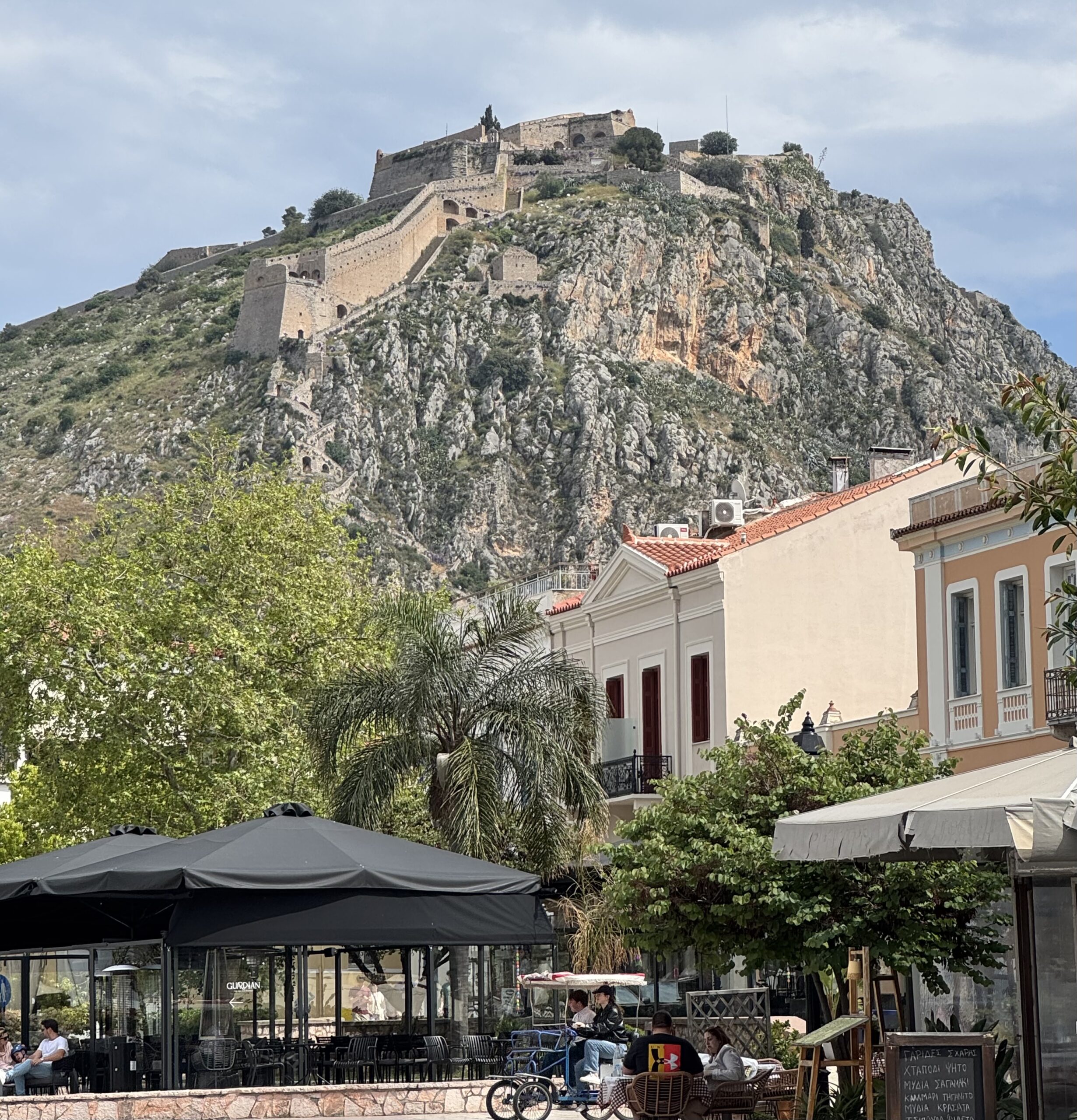IMG_8885 Palamidi - Taussig Travel Stone fortress perched on a rocky hill above a street-level cafe and outdoor seating area.