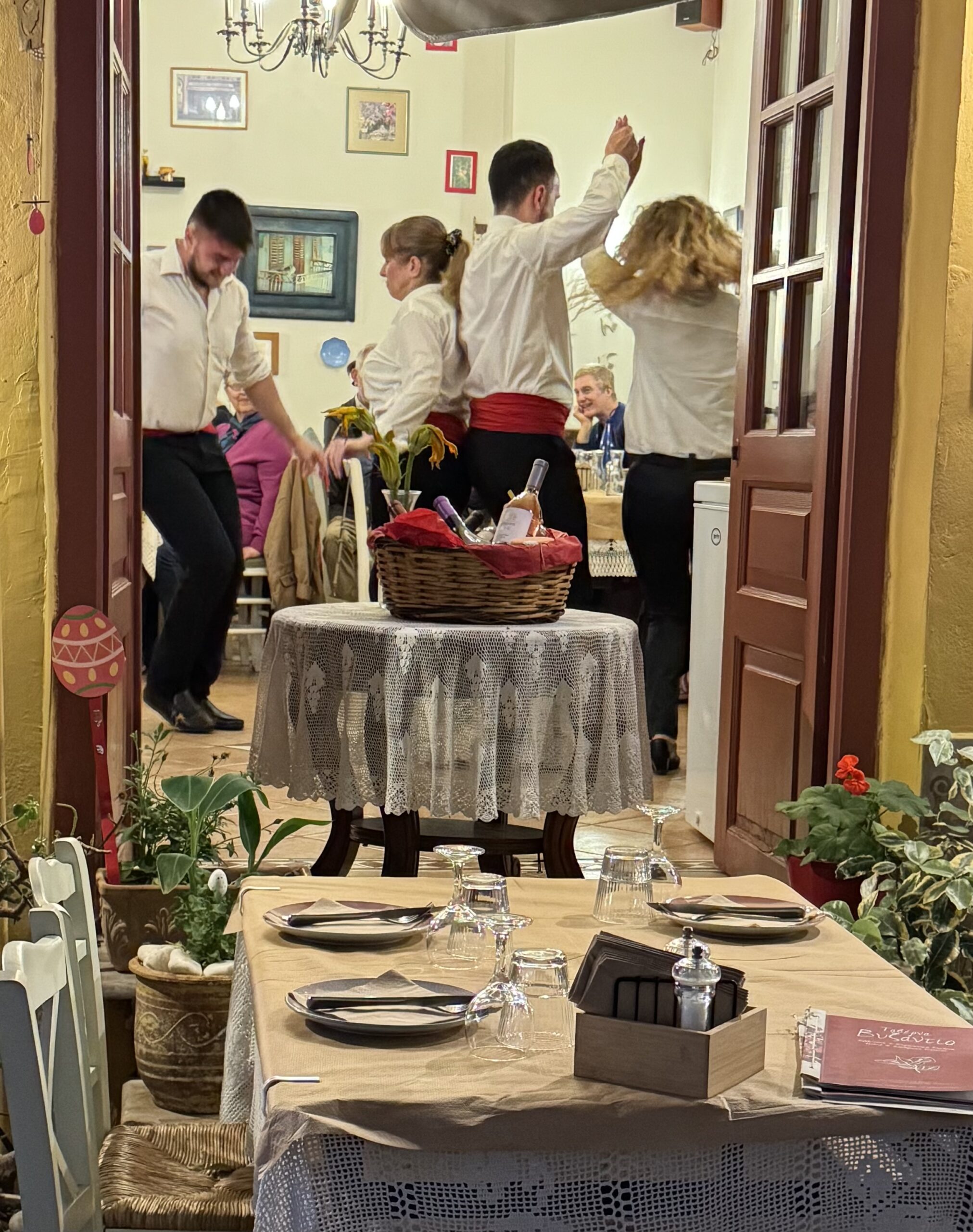 IMG_8928 Taverna - Taussig Travel Restaurant scene viewed from doorway: people in white shirts dancing, red sashes around waists, lace tablecloths and wine basket centerpiece visible inside.