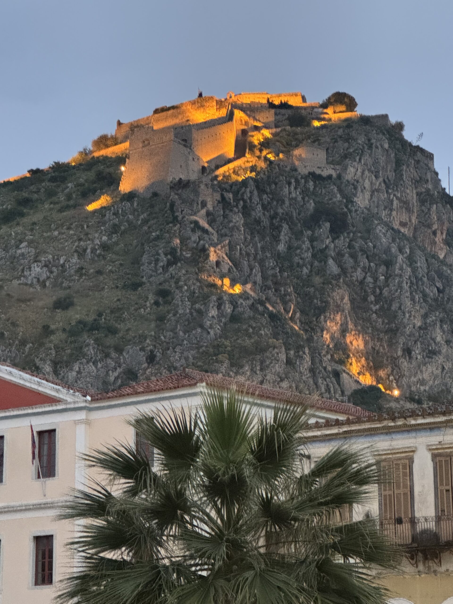 IMG_9053 Palamidi - Taussig Travel Hilltop stone fortress illuminated in warm orange lights at dusk, perched on a rocky hill above buildings with a palm tree in the foreground