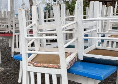 Rows of white wooden chairs stacked on blue benches in an outdoor patio. A stone wall and trees are in the background.