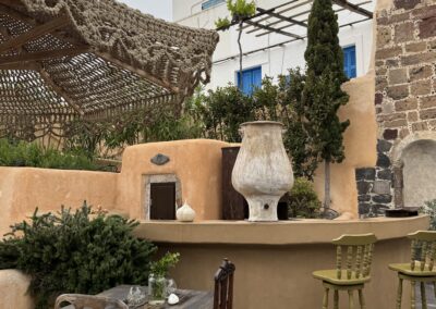 Rustic outdoor courtyard with a weathered wooden table and chairs, woven rope canopy, and orange stucco walls with blue shutters.