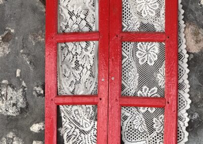 Red wooden window frame with white lace curtains mounted on a textured stone wall or exterior surface of a building.