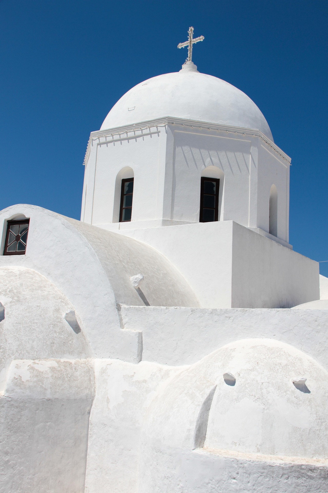 pat_photographies-megalochori-4382241_1920 - Taussig Travel White-washed church with a domed roof and cross against a bright blue sky, architectural arches and windows visible at the base of the dome