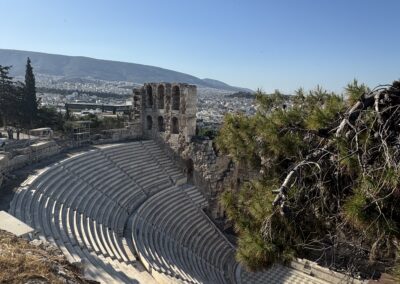 Ancient stone amphitheater with curved seating and ruined arches, overlooking a city on a hill under a clear blue sky.