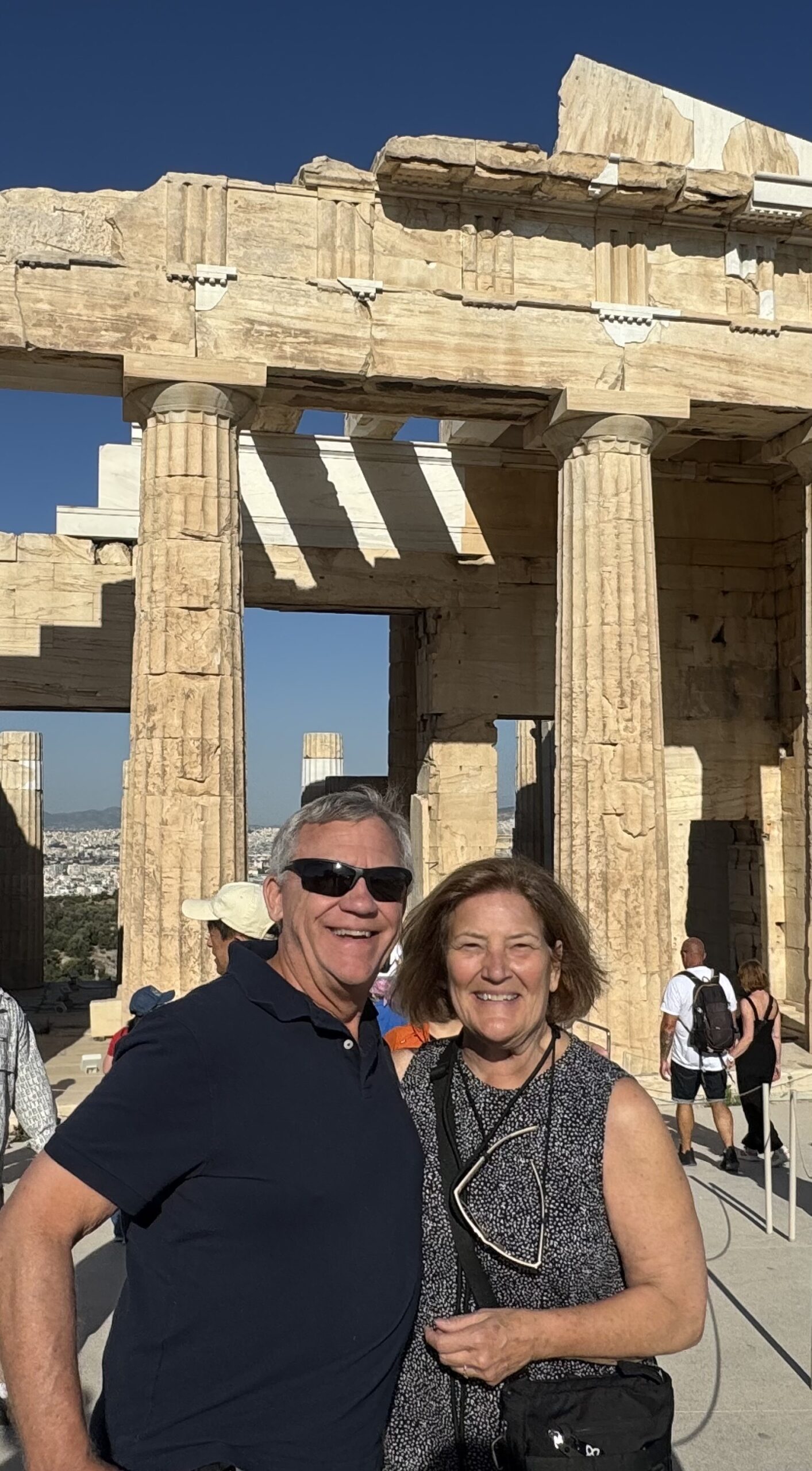 IMG_9742 Acropolis - Taussig Travel Smiling couple posing in front of ancient Greek ruins with tall limestone columns under a clear blue sky.