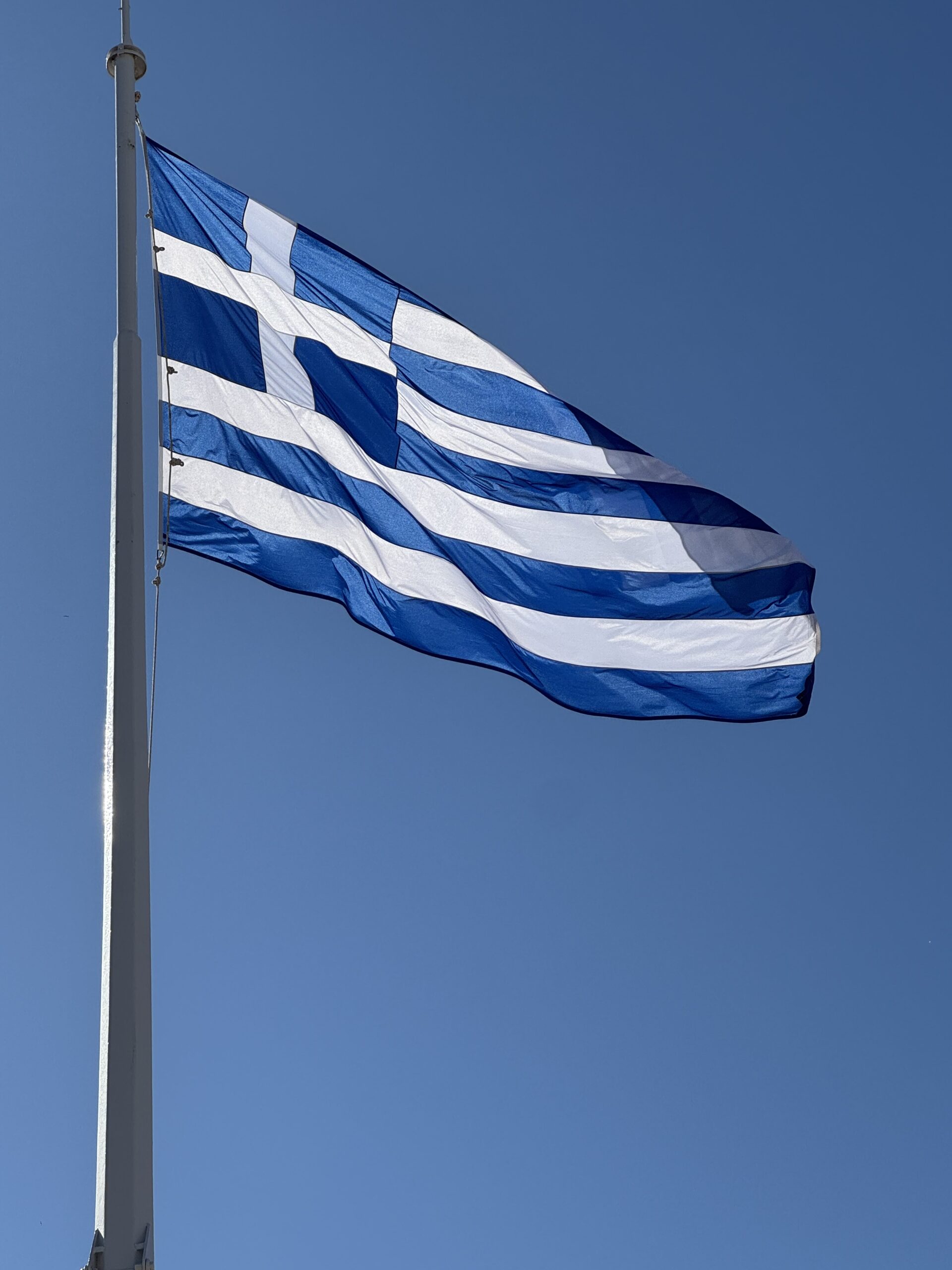IMG_9804 - Taussig Travel Flag with blue and white horizontal stripes waving on a tall pole against a clear blue sky, resembling the Greek flag.