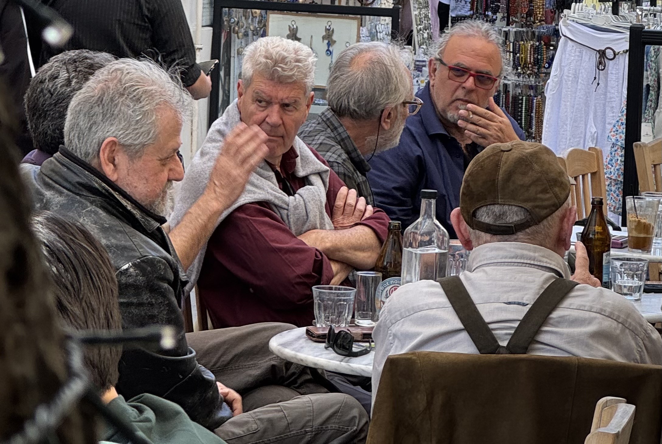IMG_9961 - Taussig Travel A group of older men seated around a round marble table at an outdoor market cafe, engaged in conversation with beer bottles and glasses on the table and market stalls visible behind them.